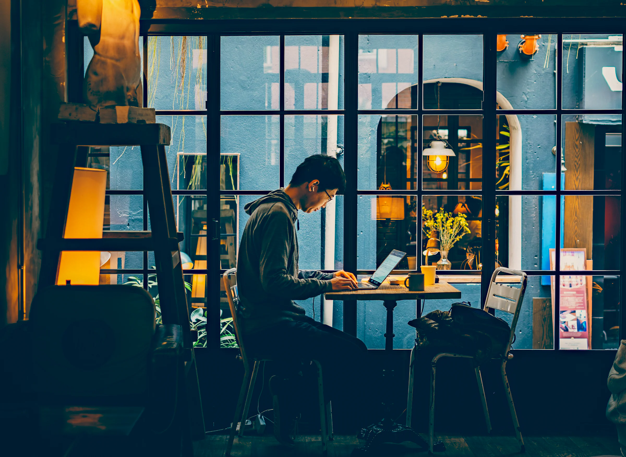 A man working on a laptop at a café table beside a tall window in the evening.