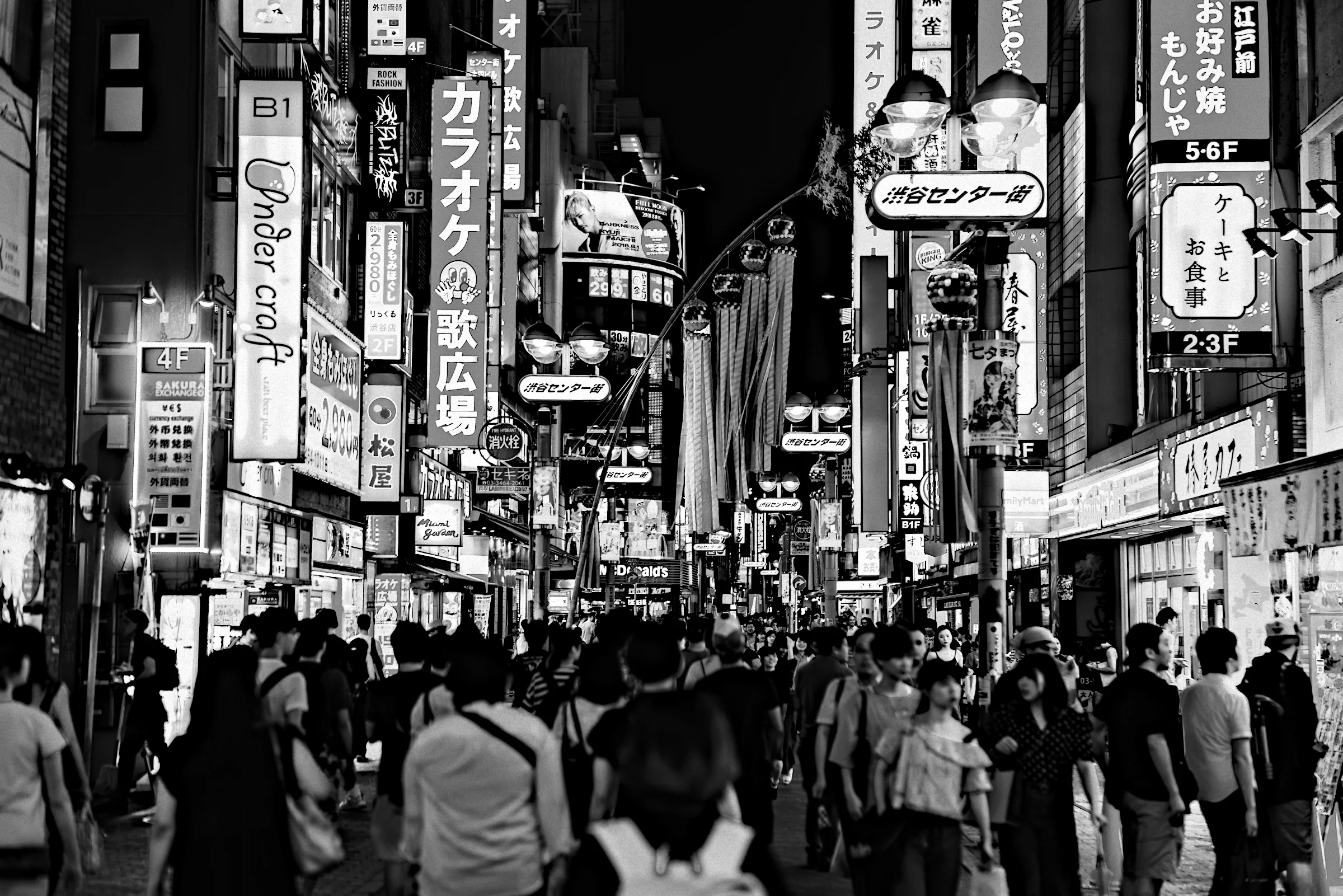 A crowd crossing a street in Tokyo at night under walls of illuminated signs, in black and white.